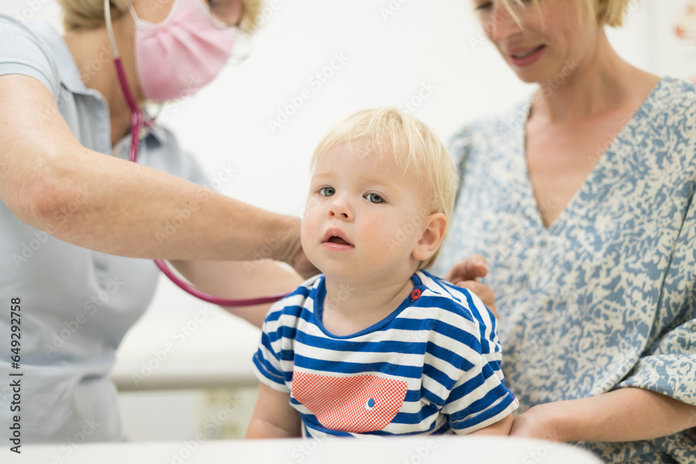 Infant baby boy child being examined by his pediatrician doctor during a standard medical checkup in presence and comfort of his mother. National public health and childs care care koncept Infant baby boy child being examined by his pediatrician doctor during a standard medical checkup in presence and comfort of his mother. National public health and childs care care koncept