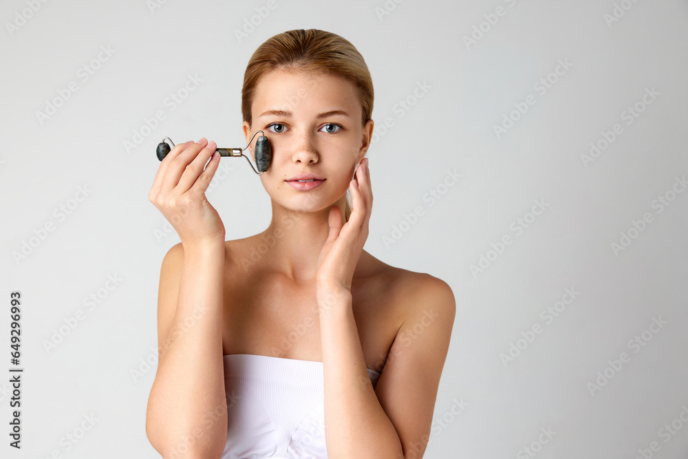 Beautiful young girl with well-kept smooth skin doing face massage with roller isolated over grey studio background