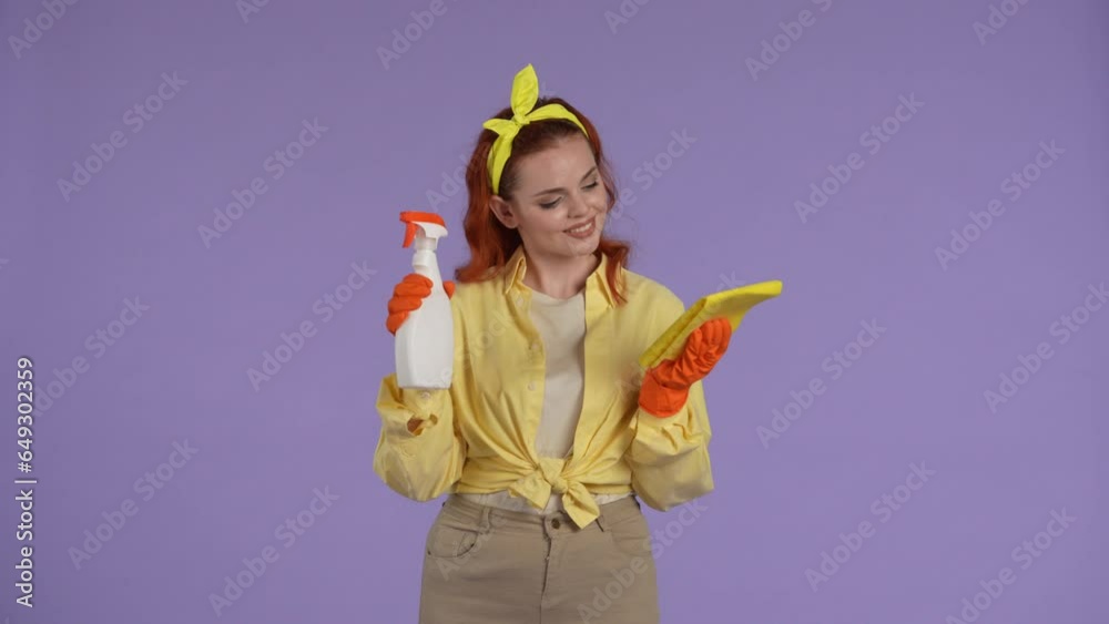 Woman in casual clothing and rubber gloves holding bottle of cleanser spray and rag, posing and smiling at the camera. Isolated on purple background. Woman in casual clothing and rubber gloves holding bottle of cleanser spray and rag, posing and smiling at the camera. Isolated on purple background.