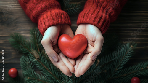 Person Holding Red Christmas Heart in Hands. Wearing Red Sweater with Pine Needle Branch in Background. Giving Concept.