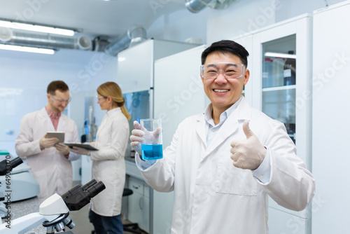 Asian male scientist, chemist, pharmacist standing in a laboratory and holding a beaker with a substance in his hands, pointing a super finger at the camera, colleagues are discussing behind.