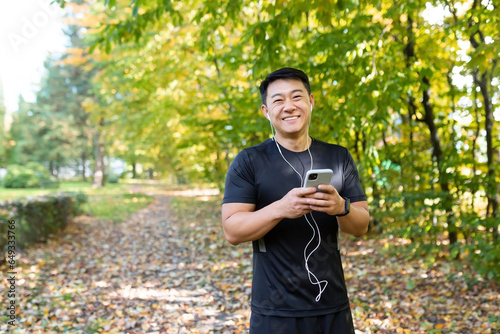 Portrait of a young Asian man running and doing sports in the park. Standing in headphones, using the phone and smiling at the camera.