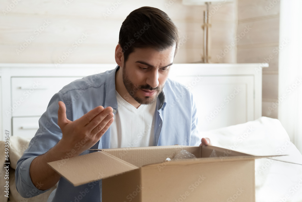 Angry irritated man unboxing parcel, looking into open cardboard box, sitting on couch at home, dissatisfied annoyed customer received wrong or broken online store order, bad delivery service concept