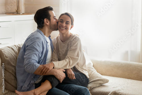 Loving boyfriend kissing smiling girlfriend on cheek, enjoying tender moment, cuddling and hugging, sitting on cozy couch in living room, happy wife and husband spending romantic date at home