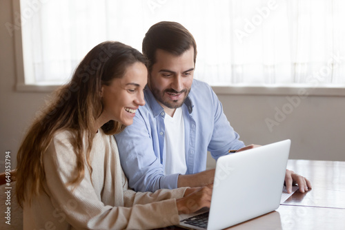 Happy young couple using laptop together, looking at screen, smiling woman and man reading good news in email, refund or loan mortgage approval, checking online banking service, shopping