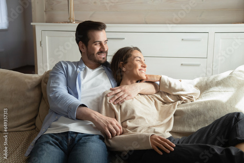 Dreamy smiling young man and woman hugging, relaxing on couch, looking to aside, happy wife and husband dreaming about good future, new opportunities, enjoying leisure time together at home