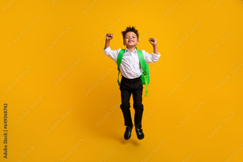 Full length photo of small beautiful schoolboy wear stylish shirt rucksack jump clenching fists isolated on yellow color background Full length photo of small beautiful schoolboy wear stylish shirt rucksack jump clenching fists isolated on yellow color background
