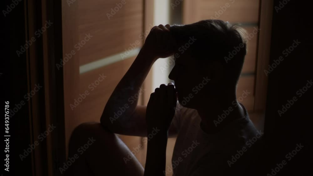 Silhouette of sad pensive man vaping e-cigarette sitting on floor in dark room. Thoughtful male in depression, calms nerves with puffs of electronic cigarette. Bad habit, addiction of smoking. Silhouette of sad pensive man vaping e-cigarette sitting on floor in dark room. Thoughtful male in depression, calms nerves with puffs of electronic cigarette. Bad habit, addiction of smoking.