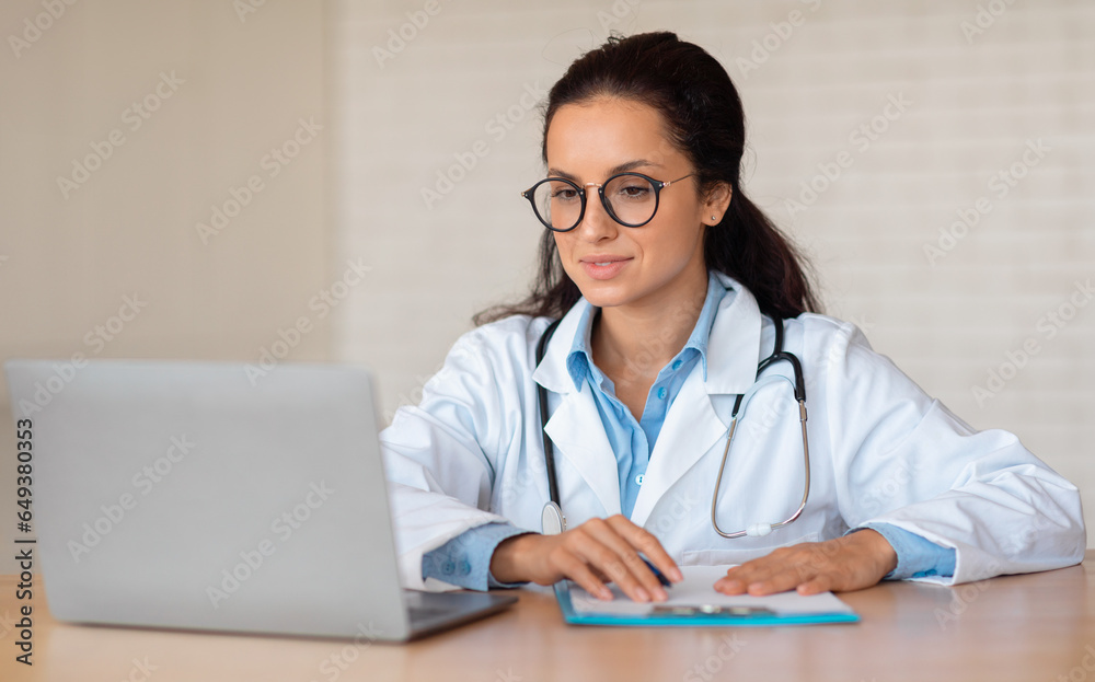 Young caucasian female nutritionist doctor in white coat making notes in clipboard, looking at laptop, sitting at table Young caucasian female nutritionist doctor in white coat making notes in clipboard, looking at laptop, sitting at table