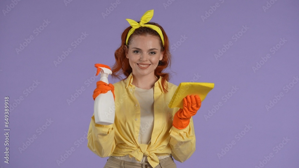 Woman in casual clothing and rubber gloves holding bottle of cleanser spray and rag, smiling. Isolated on purple background. Woman in casual clothing and rubber gloves holding bottle of cleanser spray and rag, smiling. Isolated on purple background.