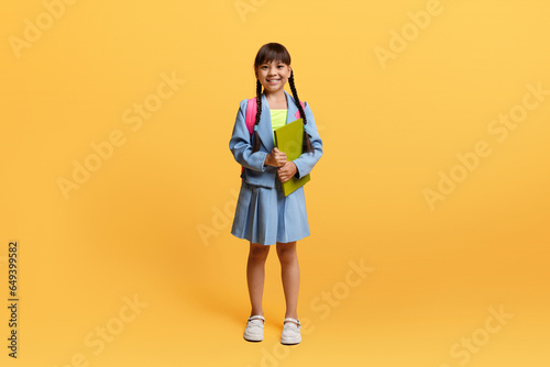 Happy asian schoolgirl with backpack and exercisebook posing on yellow photo