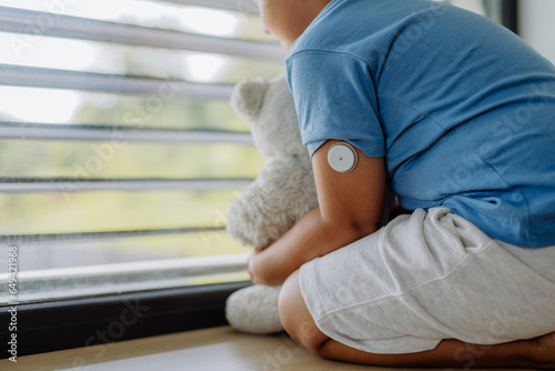 Diabetic boy with a continuous glucose monitor sitting by the window, holding his stuffed teddy bear and looking outside.