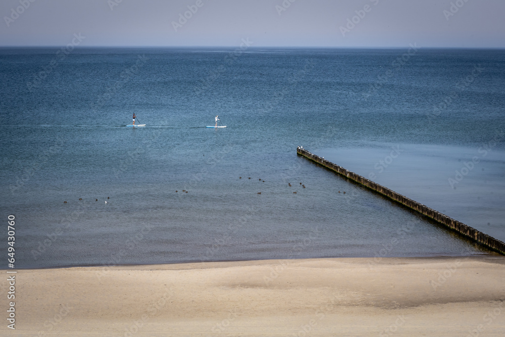 Golden sand beach with wooden breakwaters, Baltic sea, Poland. Two stand up paddle surfers on water. Golden sand beach with wooden breakwaters, Baltic sea, Poland. Two stand up paddle surfers on water.