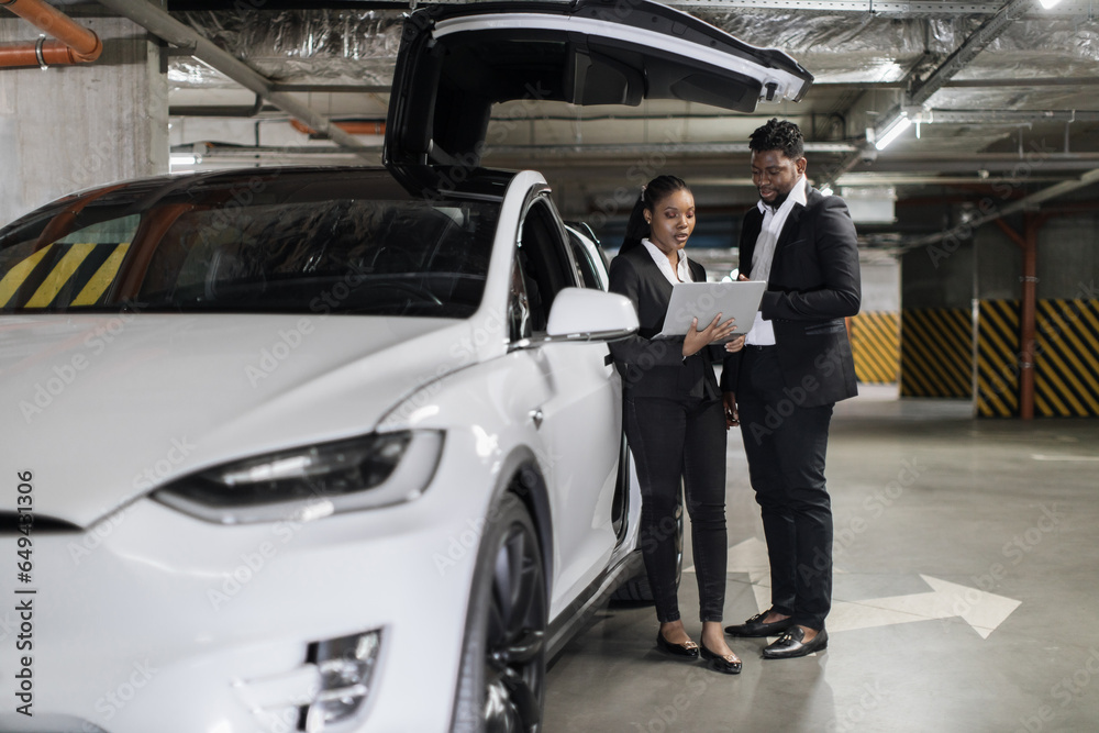 Full length view of african male and female colleagues working on laptop near open wing doors of EV in parking lot. Professional teammates discussing project strategy via device outside office walls.