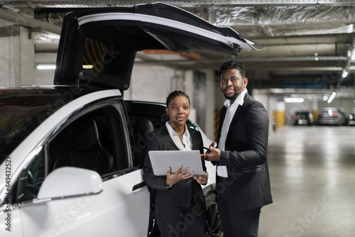 Close up view of african american adults in business suits performing tasks on portable computer by electric vehicle. Fellow colleagues working together on new project after intense hours in office.