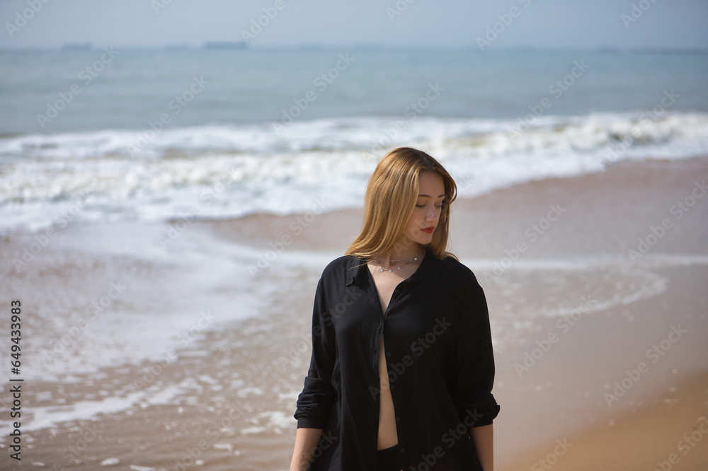 Young woman, blonde and beautiful, with a bikini and black shirt walking on the beach, relaxed, quiet, peaceful, calm. Concept of solitude, relaxation, tranquility, peace.