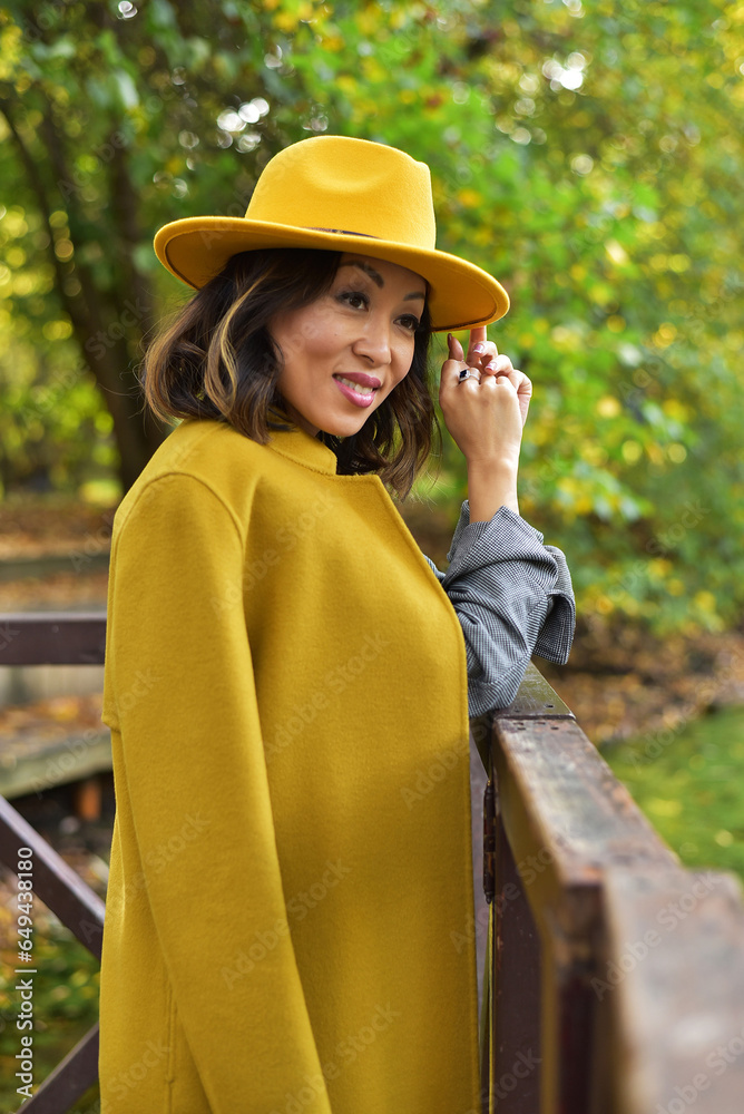 Beautiful Asian businesswoman in grey suit, yellow hat, yellow coat in the park close to to lake. Autumn.