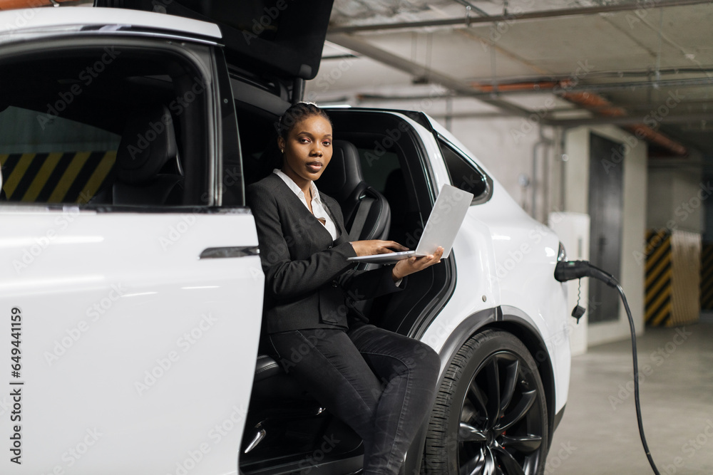 Low angle view of charming female waiting for files being downloaded on laptop while sitting on driver's seat of EV. African american employee enrolling in online programme in e-car public garage.