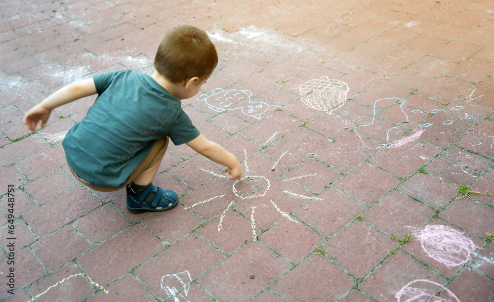 Little toddler boy two or three years old draws sun with crayons in the park on tiles. Playtime for children. Children's games. Selective focus Little toddler boy two or three years old draws sun with crayons in the park on tiles. Playtime for children. Children's games. Selective focus