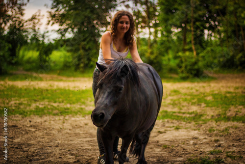 A black baby horse with hair looking at camera with a girl an out of focus girl in the background