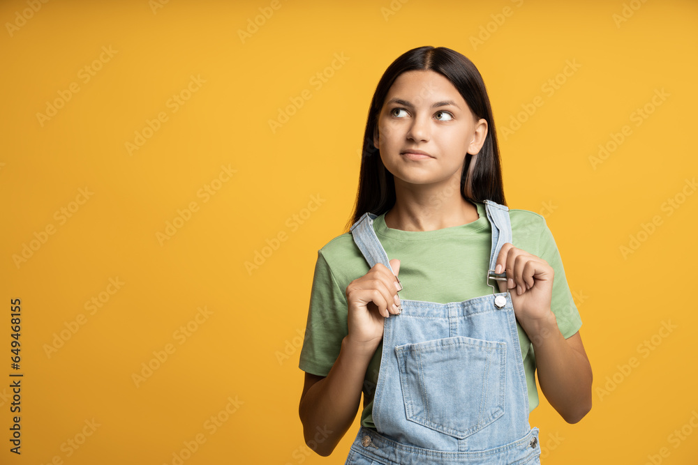 Inspired teenage girl looks away at left corner with copy space. Isolated on yellow. Young teen girl holding on to straps of denim jumpsuit with curious interested inquisitive encouraged excited look