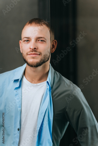 Vertical portrait of young bearded caucasian man in shirt looking at camera leaning on glass door, purposeful guy dreaming about career. Achivements and people efforts concept. photo