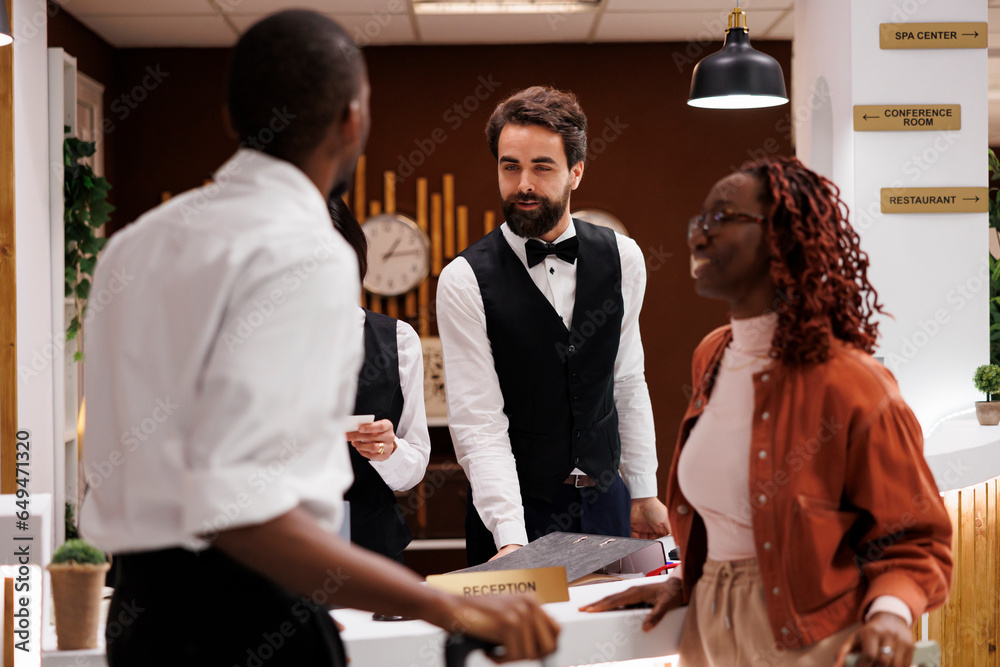 Hotel staff welcoming guests at counter, providing excellent check in service at hotel. Young employee talking to couple with luggage, looking for accommodation upon their arrival.