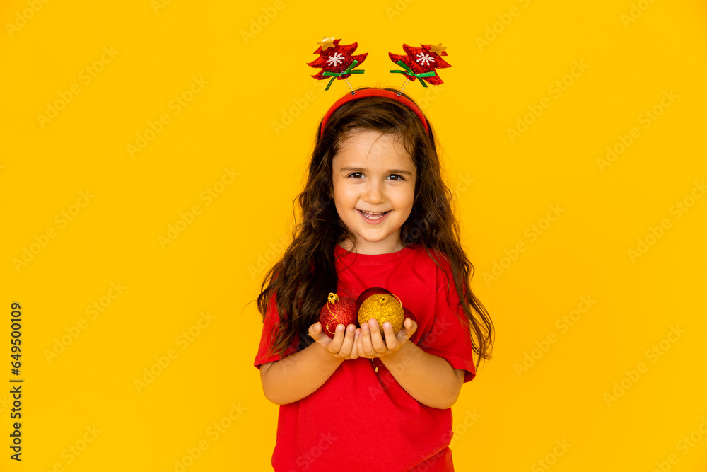 A cute little girl in a red t-shirt holding Christmas tree toys. yellow background, space for text A cute little girl in a red t-shirt holding Christmas tree toys. yellow background, space for text