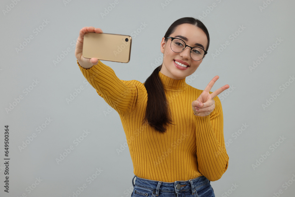 Smiling young woman taking selfie with smartphone and showing peace sign on grey background Smiling young woman taking selfie with smartphone and showing peace sign on grey background