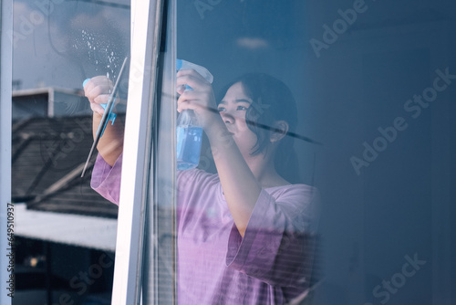 Young Asian woman cleaning window glass with rag and spray indoors