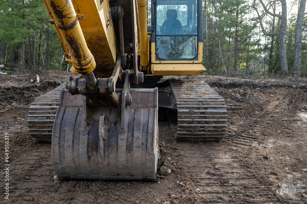 Closeup of the bucket and tracks of an excavator on the soil near trees, in a new home construction site.