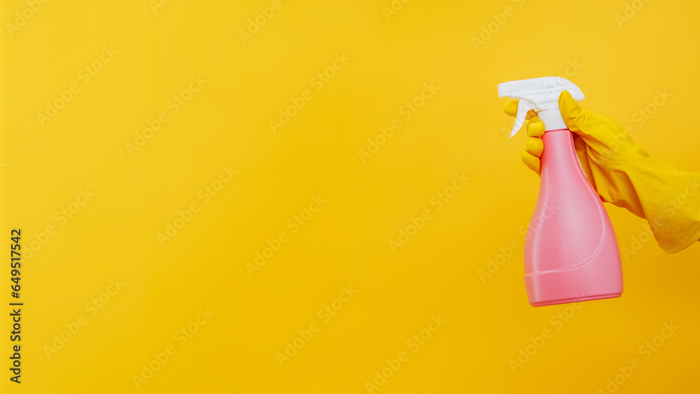 Cleaning product. Household hygiene. Home disinfection. Janitor hand in protective gloves with pink spray bottle with detergent fluid isolated on yellow background empty space. Cleaning product. Household hygiene. Home disinfection. Janitor hand in protective gloves with pink spray bottle with detergent fluid isolated on yellow background empty space.