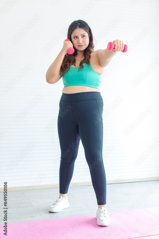 Asian young chubby fat healthy oversized overweight fit female sportswoman in casual sportswear and boxing gloves standing on yoga mat smiling punching air exercising training in home fitness gym Asian young chubby fat healthy oversized overweight fit female sportswoman in casual sportswear and boxing gloves standing on yoga mat smiling punching air exercising training in home fitness gym