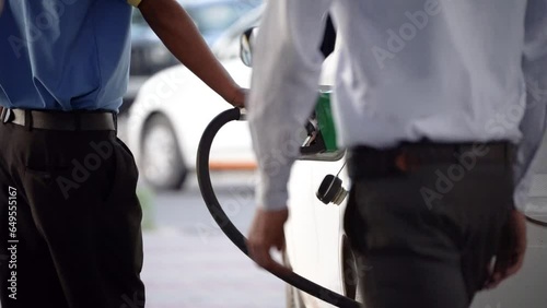 man manually filling fuel using a green nozzle into white car with people crossing showing the fuel industry in india and a major price component of Indian households