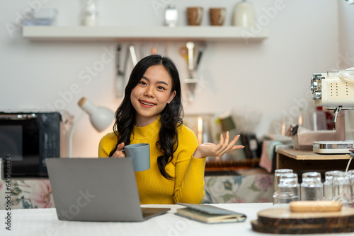 Smiling happy beautiful Asian woman relaxing using technology of laptop computer. take note of information Have a relaxing hot drink while sitting on the table in your room at home.