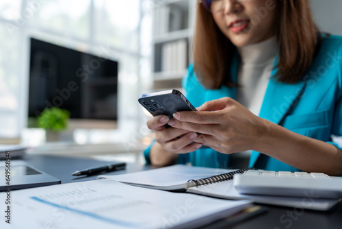 Asian businesswoman holding a smartphone using a calculator to calculate on graph paper Work record Budget calculations, taxes, financial accounts placed on the table in the office Management concept