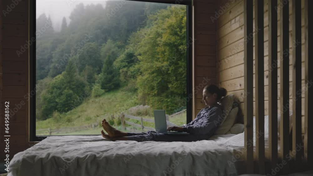 Cheerful woman working from bedroom with wide window, foggy morning in forest behind, calm leisure time concept