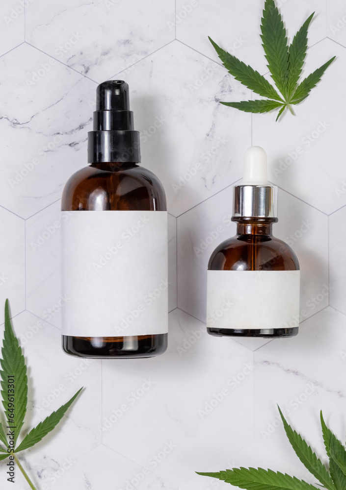 Cosmetic bottles with blank labels near green cannabis leaves on marble table. Mockup