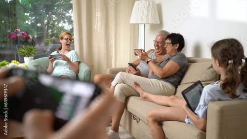 Big happy family is playing online mobile game together at home, people different ages having fun sitting on sofa. Parents, grandparents and little girl relaxing in living room with mobile devices