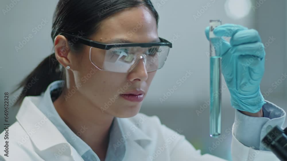 Asian female scientist in protective eyewear and gloves examining blue chemical in test tube while working in laboratory Asian female scientist in protective eyewear and gloves examining blue chemical in test tube while working in laboratory