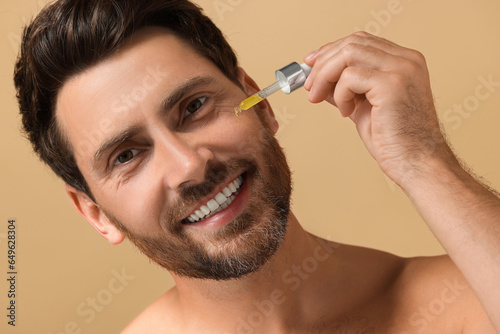 Smiling man applying cosmetic serum onto his face on beige background, closeup