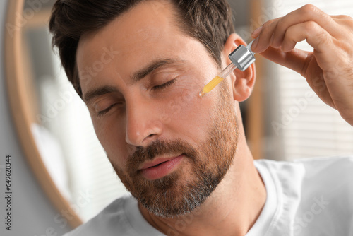 Handsome man applying cosmetic serum onto his face indoors, closeup