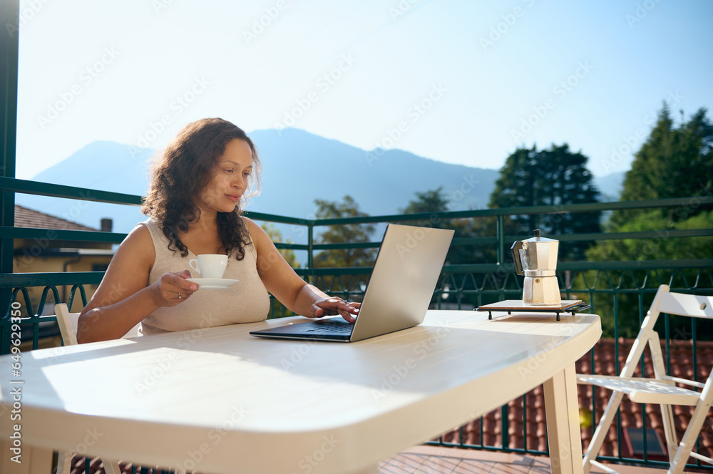Happy confident woman entrepreneur drinking coffee and using laptop checks her mail, working remotely from home terrace
