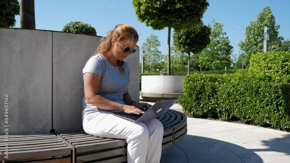 A modern young woman is working with a laptop while sitting on a round bench in a modern park among beautiful trimmed bushes. A student is engaged with a laptop in the park in the summer. A modern young woman is working with a laptop while sitting on a round bench in a modern park among beautiful trimmed bushes. A student is engaged with a laptop in the park in the summer.
