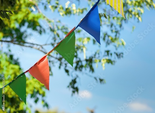 colorful pennant string decoration in green tree foliage on blue sky, summer party background. AI Generated.