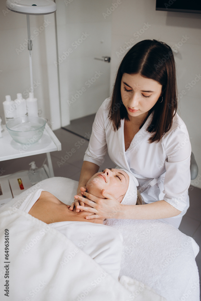Fototapeta premium Cosmetologist is wiping woman's face using paper napkins before peeling and massage procedure. Doctor beautician is making beauty procedure in cosmetology clinic. Portrait of young woman, top view.