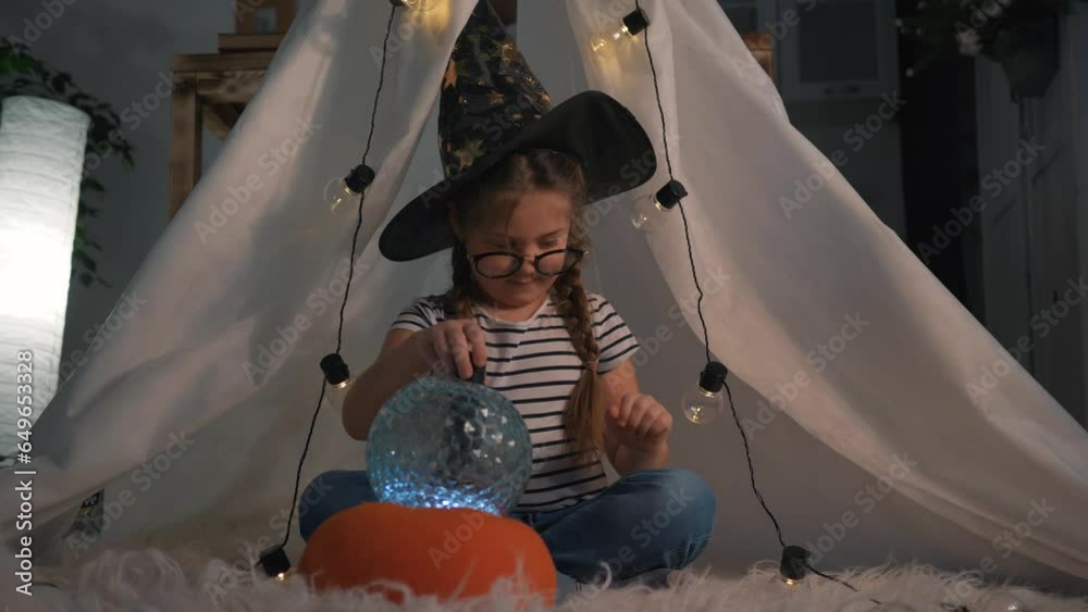 Cute happy child girl in top hat sits in tent, plays with magic lantern with glass ball.child fantasizes, dreams of witchcraft, fulfillment of desires.Happy child at home playing in room for christmas