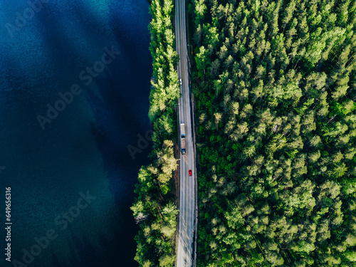 Aerial view of road with cars between green forest and blue lake in Finland