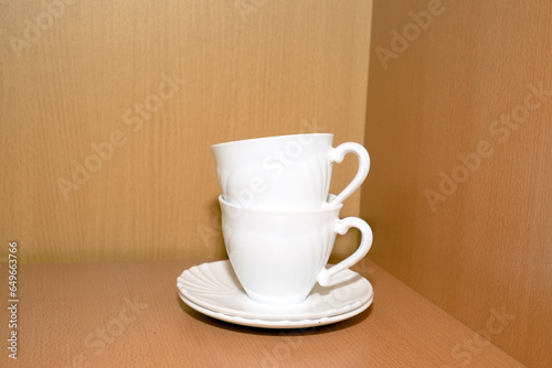 Two white mugs with saucers for tea or coffee stand in a wooden cupboard. Kitchen, tea party