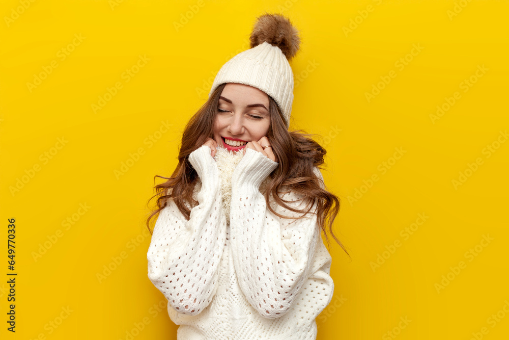 young cute girl in warm soft winter clothes smiles on yellow isolated background, woman in white hat and scarf